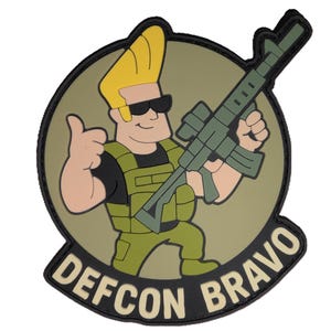 May include: A patch featuring a cartoon character with yellow hair and sunglasses, holding a green rifle and giving a thumbs-up. The character is wearing a green vest and uniform. The patch has the words "DEFCON BRAVO" in white.