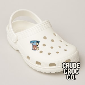 May include: White Crocs shoe with a "SWEET-T" graphic featuring a cartoon beverage. The shoe has multiple ventilation holes and a pivoting heel strap. The brand name "CRUDE CROC CO." is printed on the side.