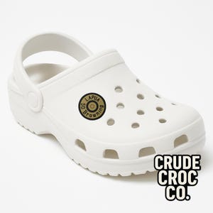 May include: White clog-style shoe with a strap and multiple ventilation holes. A circular emblem with the text "LAPUA BROWNING 50" is on the side. The text "CRUDE CROC CO." is printed in a block font on the side.