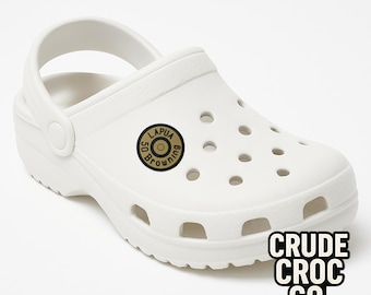 May include: White clog-style shoe with a strap and multiple ventilation holes. A circular emblem with the text "LAPUA BROWNING 50" is on the side. The text "CRUDE CROC CO." is printed in a block font on the side.