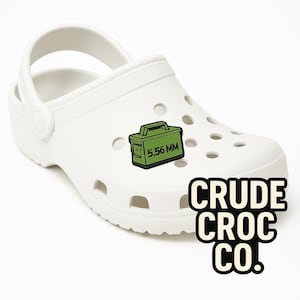 May include: White Crocs shoe with a green and black 5.56 MM ammo box charm. The shoe has multiple holes and a pivoting heel strap. The words "CRUDE CROC CO." are printed in black and white.