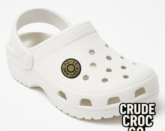 May include: White clog-style shoe with a pivoting heel strap. Features a circular emblem with "9MM LUGER" text and "FC 20" in the center. The text "CRUDE CROC CO." is printed in a black and white block font on the side.