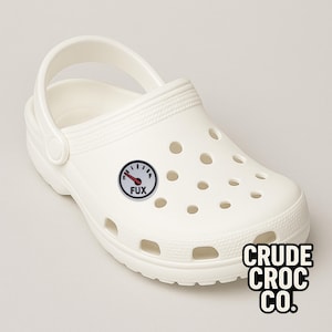May include: White clog shoe with a black and white gauge charm that reads "FUX". The charm is round with a red needle. The shoe has multiple holes and a pivoting heel strap. The text "CRUDE CROC CO." is printed on the side.