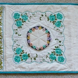 May include: A white quilted table runner with three square panels featuring vintage handkerchiefs with floral and geometric designs. The center panel has a crocheted circle in the center.