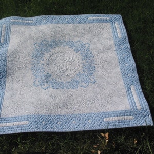 May include: A white quilt with a blue floral design in the center and a blue border. The quilt is lying on a green grassy surface.
