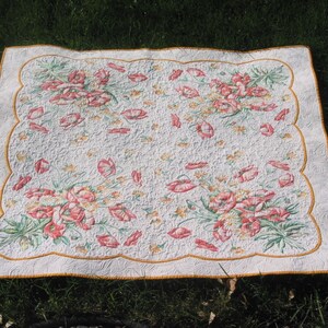 May include: A white quilt with a scalloped border and a floral pattern featuring pink and green flowers. The quilt is quilted with a swirling pattern.