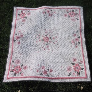 May include: A white quilted throw blanket with a pink floral pattern. The blanket has a pink and white border.