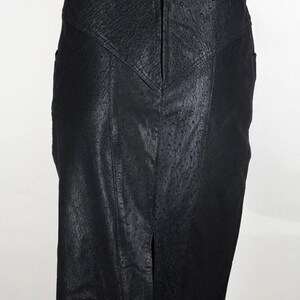 May include: A black pencil skirt with a textured, leather-like appearance. The skirt features a front slit and a zipper closure at the back. The design includes a V-shaped yoke at the waist and vertical panels.