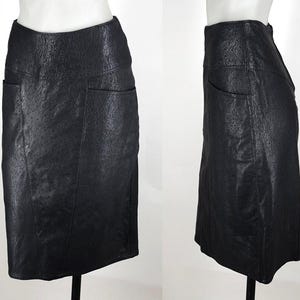 May include: A black, knee-length skirt with a textured surface and two front pockets. The skirt is displayed on a mannequin, showcasing its tailored fit and design. The skirt's material appears to be leather or a leather-like fabric.
