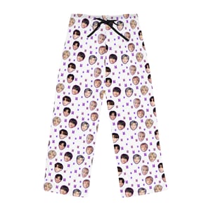 May include: White pajama pants with a repeating pattern of faces and purple symbols. The pants have a black drawstring at the waist. The faces are of various people. The pajama pants are made of a soft material.