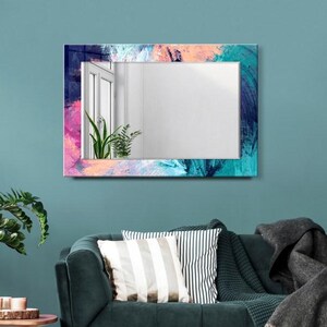 Pode incluir: Espelho de parede retangular com uma moldura pintada abstrata colorida em tons de azul, rosa e verde-azulado. O espelho está pendurado acima de um sofá verde escuro com almofadas decorativas e um cobertor cinza. Uma pequena mesa lateral de madeira e uma planta em vaso estão em primeiro plano.
