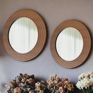 Könnte beinhalten: Zwei runde Holzspiegel mit gerilltem Design an den Rändern sind an einer grauen Wand befestigt. Die Spiegel spiegeln einen Blick auf ein Fenster wider. Getrocknete Blumen und eine Lampe sind darunter zu sehen.