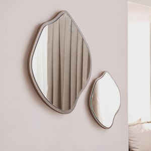 May include: Two modern, organic-shaped mirrors with a light brown frame are mounted on a pale pink wall. The larger mirror is approximately 30 inches tall, and the smaller mirror is about 18 inches tall. The mirrors reflect a neutral-toned curtain.