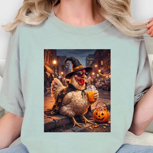 May include: A light sage green t-shirt featuring a graphic print of a chicken wearing a black witch hat, a black cape with orange pumpkin patterns, and sunglasses. The chicken holds an orange drink and stands next to a jack-o'-lantern bucket.