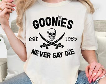 May include: An ivory short-sleeve t-shirt featuring a black graphic print. The design includes a central skull above two crossed swords, with the text GOONIES, est 1985, and NEVER SAY DIE arranged around the central imagery in a distressed font.