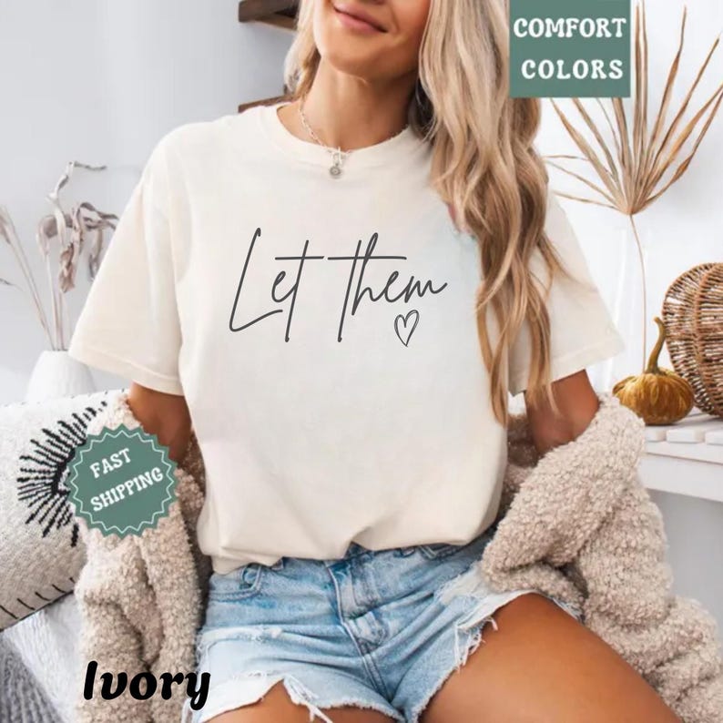 A woman wearing a white t-shirt with the text "Let Them" and a heart symbol, paired with denim shorts and a beige cardigan. She is smiling and appears to be in a relaxed setting with plants and other decorative items in the background.