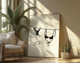 May include: A framed art print featuring a black and white illustration of a bikini hanging on a clothesline. The text "suns out, buns out!" is below the bikini. The artwork is displayed in a well-lit room with a wicker chair and plants.