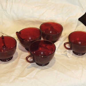 May include: Five red glass cups with a rounded bottom and a flared lip. The cups are arranged on a white tablecloth.