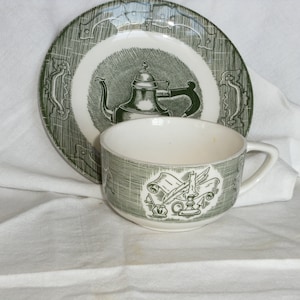 Puede incluir: Un juego de taza y platillo vintage verde y blanco. El plato presenta una ilustración de tetera, mientras que la taza muestra un diseño de pluma y pergamino. El juego es de cerámica y es adecuado para servir té.