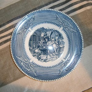 May include: Blue and white decorative plate with a farm scene in the center. The plate has a detailed border design and a rim with a beaded pattern. The farm scene includes a horse-drawn carriage, people, and farm animals.