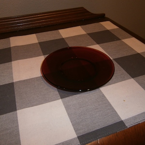 May include: A round, dark red glass plate sits on a checkered gray and white tablecloth. The plate is centered and appears to be empty. The table has a dark brown wooden edge.