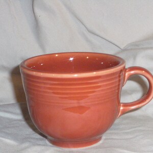 May include: A terracotta-colored ceramic mug with a rounded shape and a single handle. The mug has a smooth, glossy finish and is decorated with a series of horizontal lines around the body.