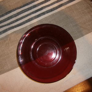 May include: A round, deep red glass saucer with a glossy finish. The saucer has a slightly raised center and a smooth, curved rim. It sits on a beige and white striped surface, suggesting a vintage or antique aesthetic.