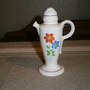 May include: White ceramic teapot with a floral design. The teapot has a spout, a handle, and a lid with holes. The flowers are orange, blue, and green.