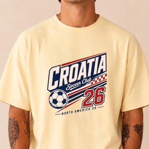 K&ouml;nnte beinhalten: Ein hellgelbes Kurzarm-T-Shirt mit einem Grafikdruck, der CROATIA Soccer Club 26 North America 26 zeigt. Das Design umfasst einen Fu&szlig;ball, ein rot-wei&szlig;es Karomuster sowie blauen und roten Text auf einem hellgelben Hintergrund.