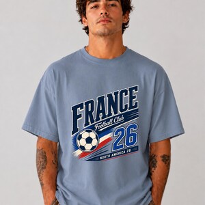 May include: A dusty blue short-sleeve t-shirt featuring a graphic print with the text FRANCE, Football Club, 26, and NORTH AMERICA 26. The design includes a football and red, white, and blue brushstroke accents across the centre of the chest.