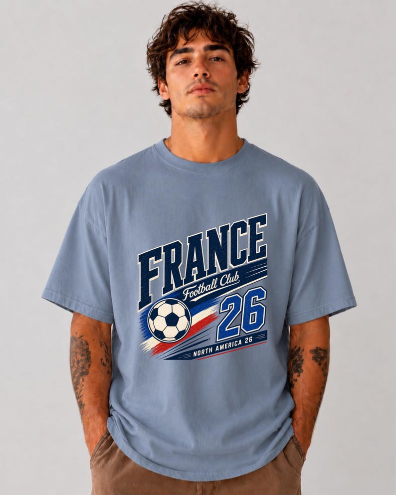 May include: A dusty blue short-sleeve t-shirt featuring a graphic print with the text FRANCE, Football Club, 26, and NORTH AMERICA 26. The design includes a football and red, white, and blue brushstroke accents across the centre of the chest.