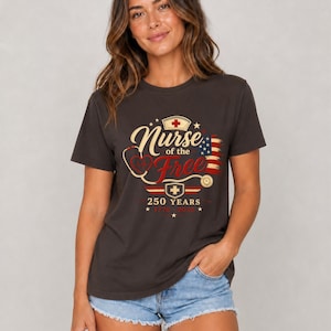May include: Dark gray t-shirt with the text "Nurse of the Free" in a stylized font, featuring a stethoscope, a nurse's cap, and an American flag design. The shirt also includes the text "250 YEARS".