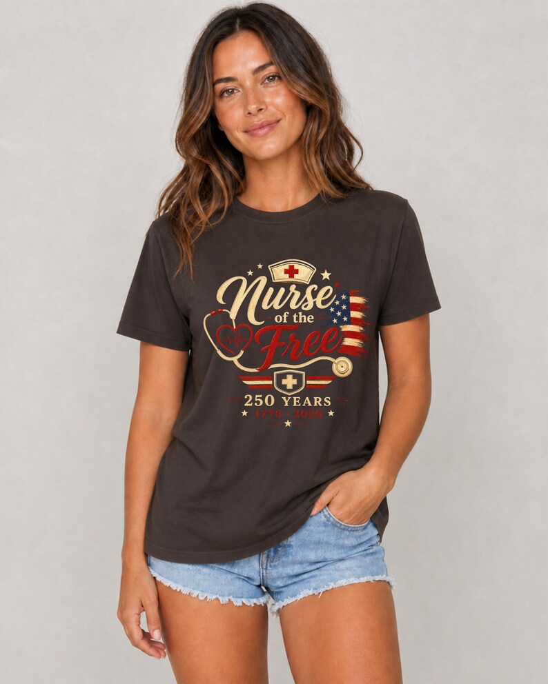 May include: Dark gray t-shirt with the text "Nurse of the Free" in a stylized font, featuring a stethoscope, a nurse's cap, and an American flag design. The shirt also includes the text "250 YEARS".