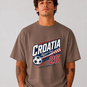 K&ouml;nnte beinhalten: Ein braunes Kurzarm-T-Shirt mit einem Grafikdruck, der "CROATIA Soccer Club" zeigt, zusammen mit einem Fu&szlig;ball, einem rot-wei&szlig;en Karomuster, der Zahl "26" und dem Text "NORTH AMERICA 26" in den Farben Blau, Rot und Wei&szlig;.