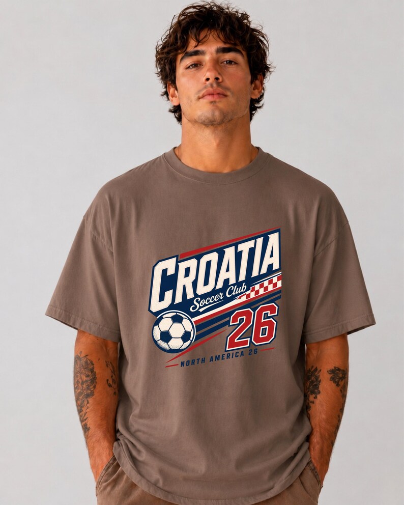 K&ouml;nnte beinhalten: Ein braunes Kurzarm-T-Shirt mit einem Grafikdruck, der "CROATIA Soccer Club" zeigt, zusammen mit einem Fu&szlig;ball, einem rot-wei&szlig;en Karomuster, der Zahl "26" und dem Text "NORTH AMERICA 26" in den Farben Blau, Rot und Wei&szlig;.