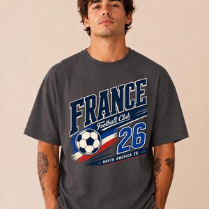 May include: A charcoal grey short-sleeve t-shirt featuring a graphic print that reads FRANCE Football Club 26 NORTH AMERICA 26. The design includes a football and blue, white, and red brushstroke accents across the centre of the chest.