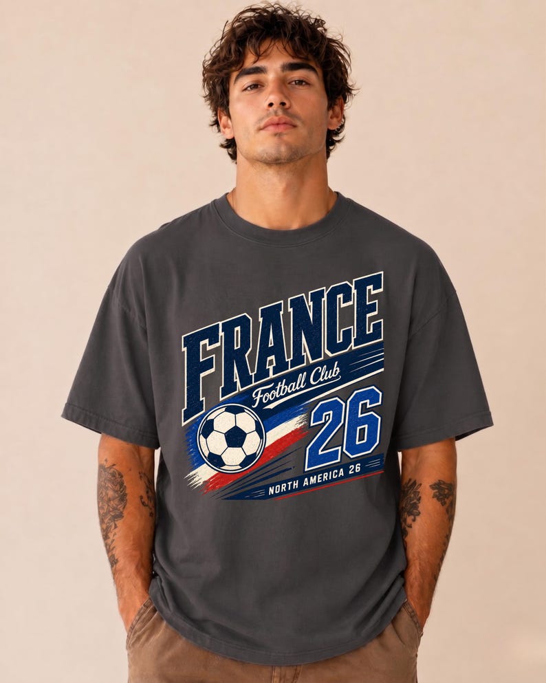 May include: A charcoal grey short-sleeve t-shirt featuring a graphic print that reads FRANCE Football Club 26 NORTH AMERICA 26. The design includes a football and blue, white, and red brushstroke accents across the centre of the chest.