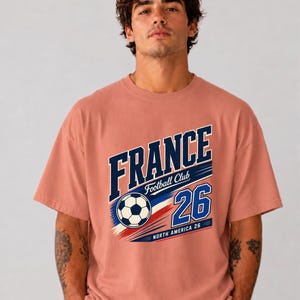 May include: A dusty rose short-sleeve t-shirt featuring a graphic print with the text FRANCE Football Club 26 NORTH AMERICA 26 and a football illustration in navy blue, white, and red colours centred on the chest area of the garment.