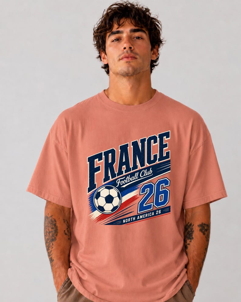May include: A dusty rose short-sleeve t-shirt featuring a graphic print with the text FRANCE Football Club 26 NORTH AMERICA 26 and a football illustration in navy blue, white, and red colours centred on the chest area of the garment.