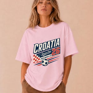 以下が含まれることがあります： CROATIA Soccer Club 26 NORTH AMERICA 26と書かれたグラフィックプリントが施されたライトピンクの半袖Tシャツ。デザインには、赤と白の市松模様の正方形、サッカーボール、赤と青の水平な動きの線が含まれています。