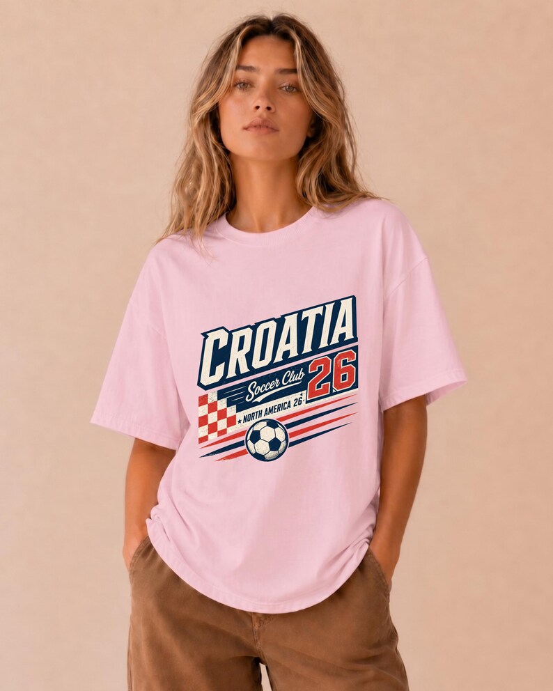 以下が含まれることがあります： CROATIA Soccer Club 26 NORTH AMERICA 26と書かれたグラフィックプリントが施されたライトピンクの半袖Tシャツ。デザインには、赤と白の市松模様の正方形、サッカーボール、赤と青の水平な動きの線が含まれています。