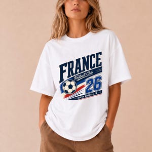 May include: A white short-sleeved t-shirt featuring a graphic print with the text FRANCE, Football Club, and the number 26. The design includes a football and red, white, and blue stripes with the additional text North America 26.