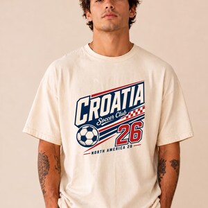 K&ouml;nnte beinhalten: Ein cremefarbenes Kurzarm-T-Shirt mit einem Grafikdruck, der "CROATIA Soccer Club" in Blau und Rot zeigt, zusammen mit einem Fu&szlig;ball, einem rot-wei&szlig;en Karomuster, der Zahl "26" und dem darunter stehenden Text "NORTH AMERICA 26".