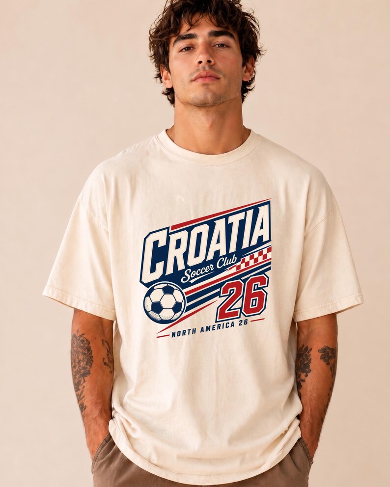 K&ouml;nnte beinhalten: Ein cremefarbenes Kurzarm-T-Shirt mit einem Grafikdruck, der "CROATIA Soccer Club" in Blau und Rot zeigt, zusammen mit einem Fu&szlig;ball, einem rot-wei&szlig;en Karomuster, der Zahl "26" und dem darunter stehenden Text "NORTH AMERICA 26".