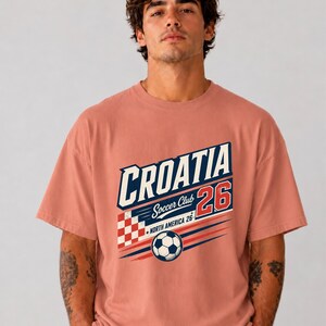 以下が含まれることがあります： CROATIA、Soccer Club、NORTH AMERICA 26という文字がデザインされた、ダスティローズ色の半袖Tシャツ。プリントには赤と白の市松模様、赤色の数字26、そして黒と白のサッカーボールが描かれています。