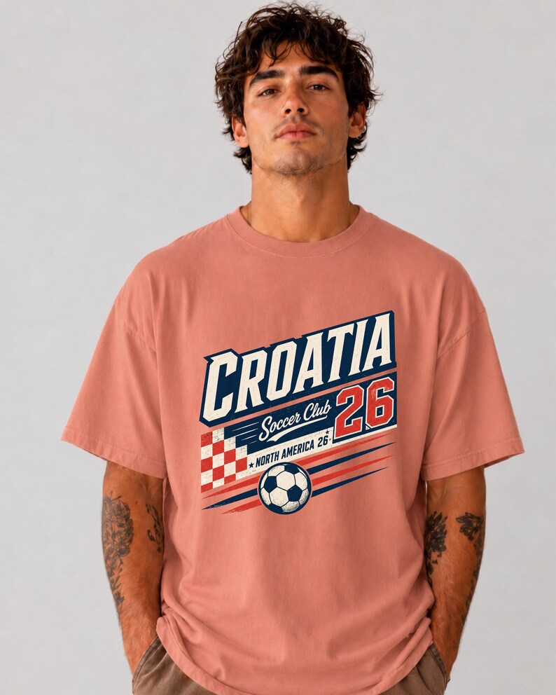 以下が含まれることがあります： CROATIA、Soccer Club、NORTH AMERICA 26という文字がデザインされた、ダスティローズ色の半袖Tシャツ。プリントには赤と白の市松模様、赤色の数字26、そして黒と白のサッカーボールが描かれています。