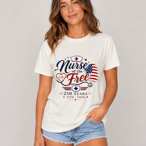 May include: Cream-colored t-shirt featuring the text "Nurse of the Free" in navy blue script, with a red heart and stethoscope design. The design includes an American flag and the text "250 YEARS 1776-2026". The model is wearing denim shorts.