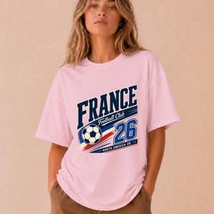 May include: A light pink short-sleeve t-shirt featuring a graphic print with the text FRANCE Football Club 26 and NORTH AMERICA 26. The design includes a football and blue, white, and red diagonal stripes across the centre of the chest.