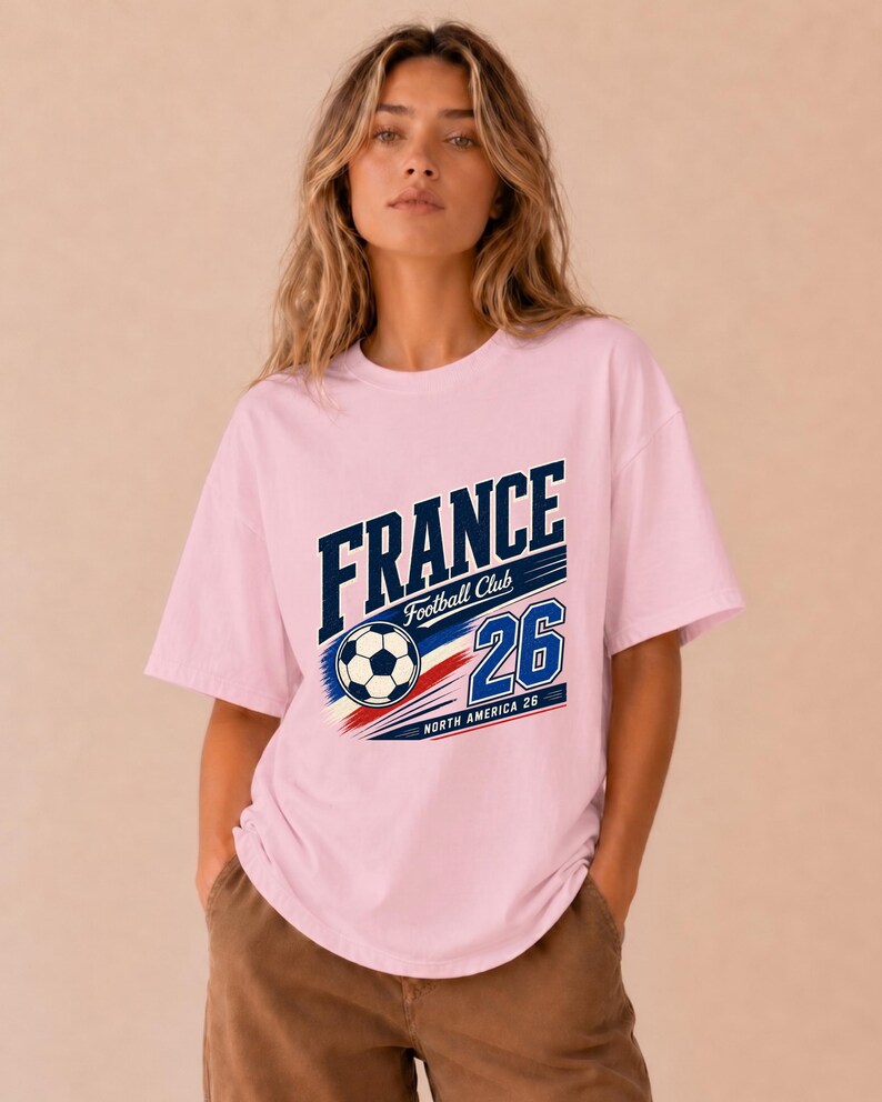May include: A light pink short-sleeve t-shirt featuring a graphic print with the text FRANCE Football Club 26 and NORTH AMERICA 26. The design includes a football and blue, white, and red diagonal stripes across the centre of the chest.