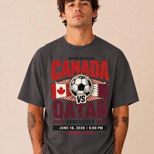 May include: Dark gray t-shirt with a distressed graphic featuring "CANADA vs QATAR" with a soccer ball, flags, and event details. The text includes "VANCOUVER JUNE 18, 2026 | 6:00 PM".